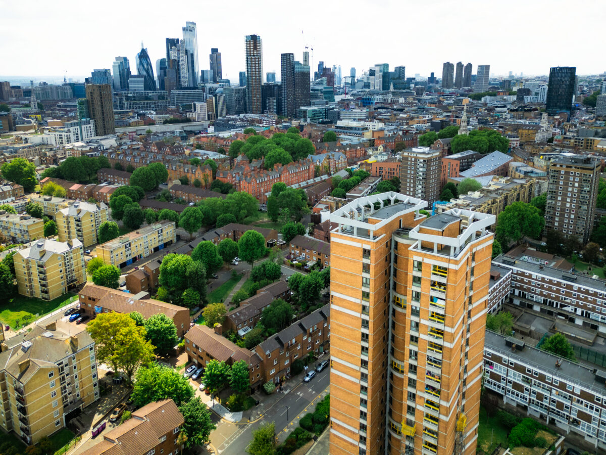 Aerial view, taken by drone, depicting the urban residential streets and apartment buildings of Hoxton, a vibrant area in Hackney, London, known for its blend of history and contemporary culture, especially in the East End and Shoreditch. On the horizon we can see the modern skyline and architecture of the city.